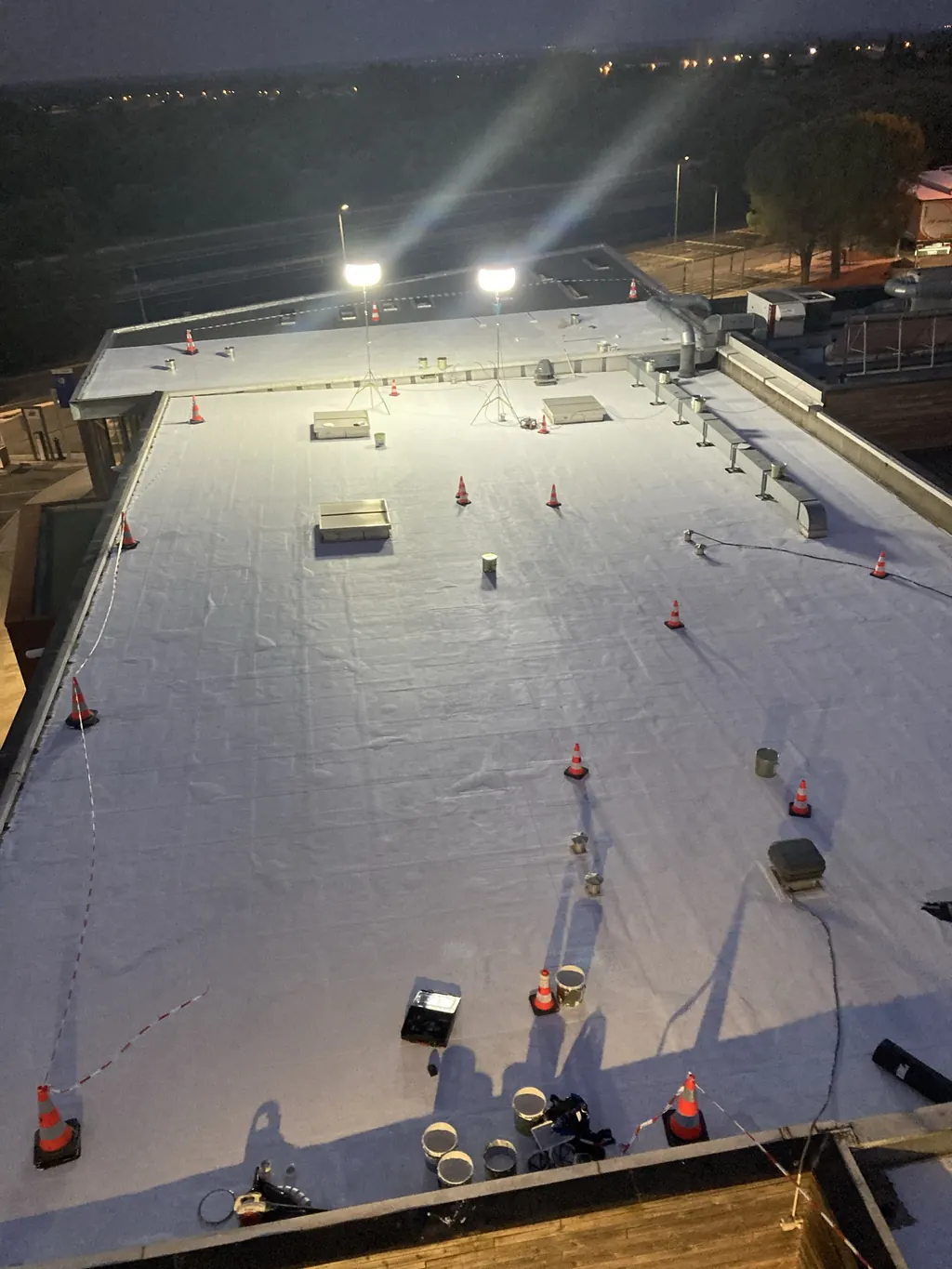 Toiture-terrasse blanche éclairée par des projecteurs et balisée de cônes, pendant un chantier de nuit